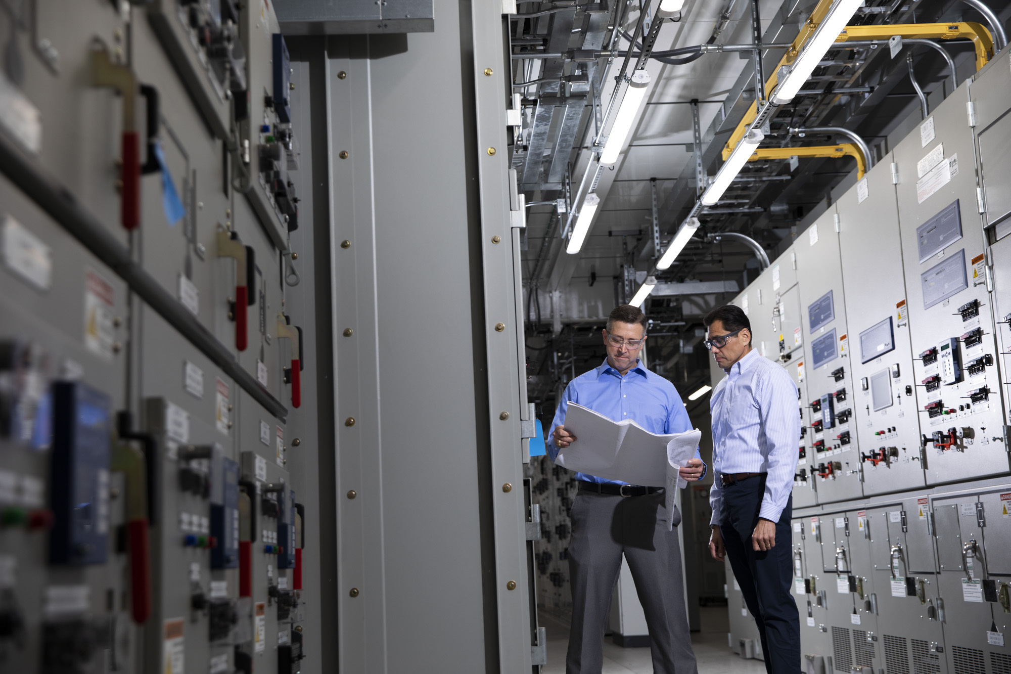 Two Powell personnel review drawings between rows of electrical switchgear