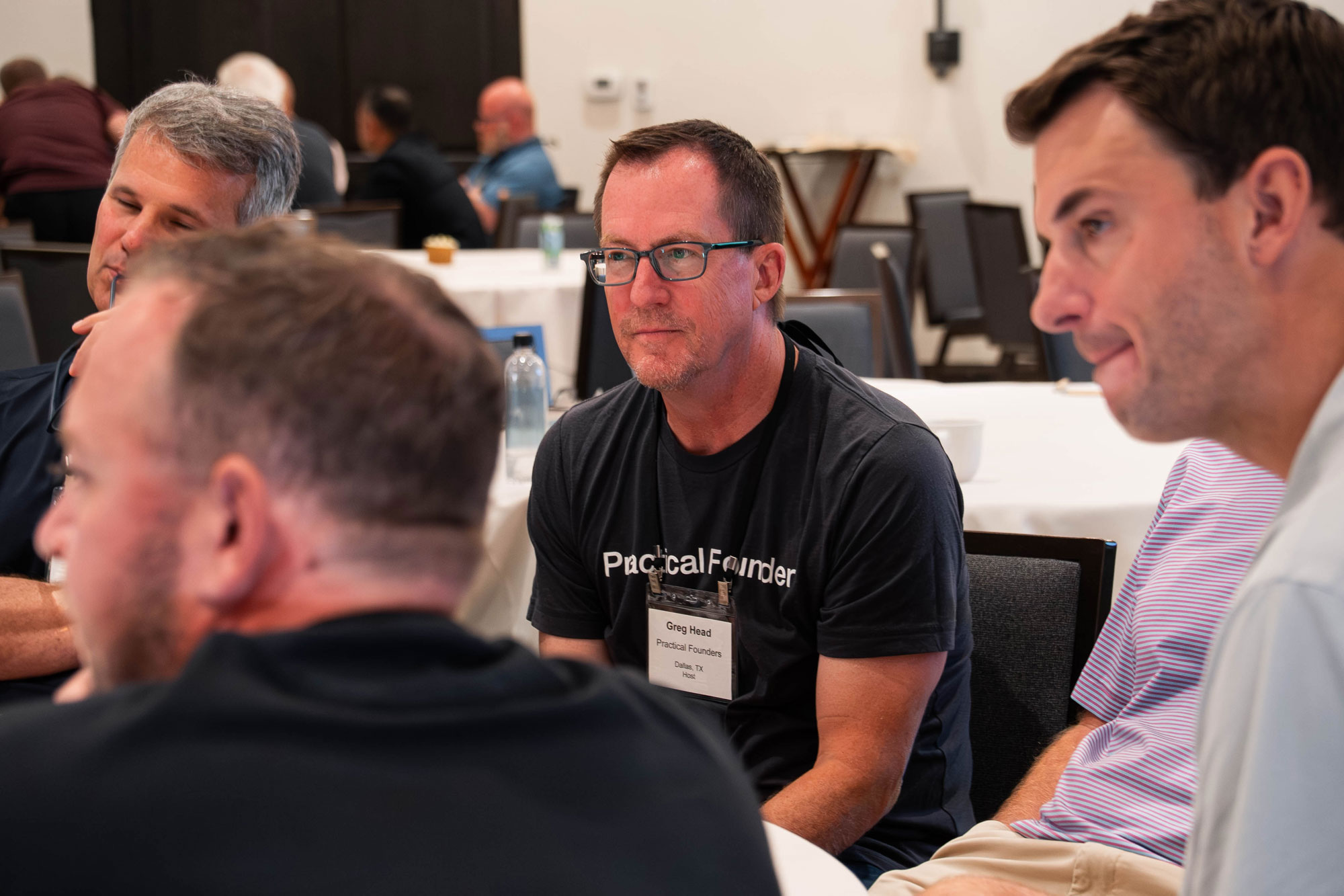 Greg Head, wearing glasses and a Practical Founders shirt, listening during a group discussion