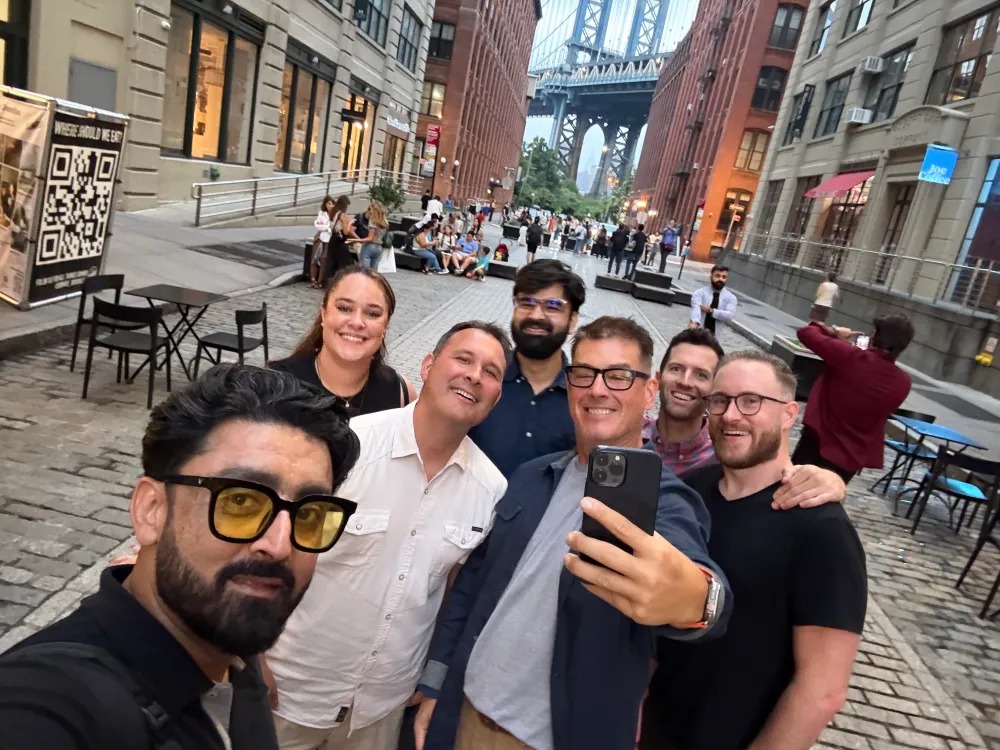 Seven Privado team members taking a group selfie on a cobblestone street near the Manhattan Bridge; Prashant Mahajan stands in the center rear wearing a dark blue shirt.