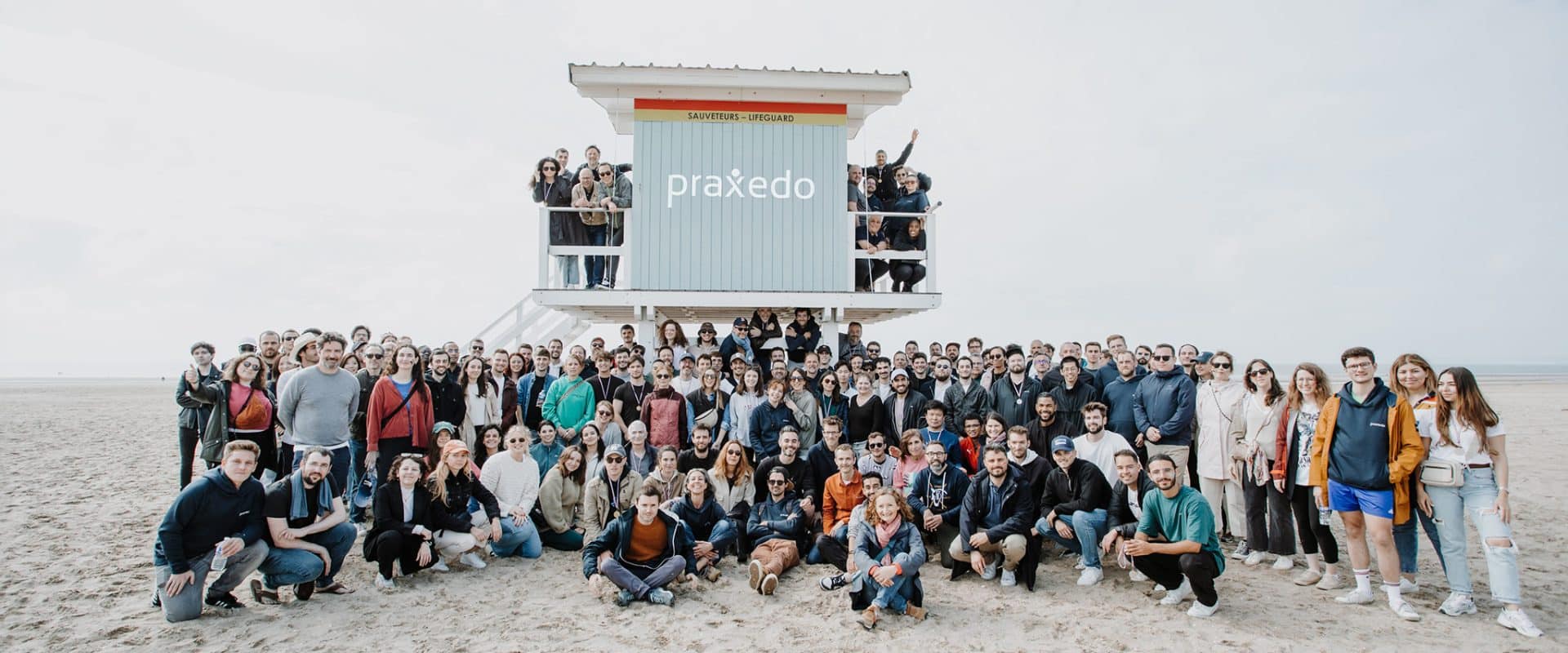A large Praxedo group gathered on a beach around a lifeguard hut
