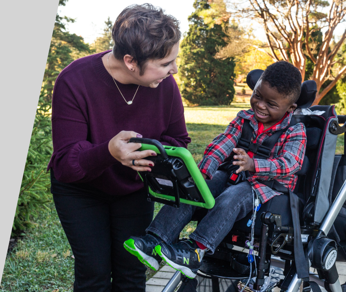 A woman holds a green communication device beside a smiling child in a wheelchair outdoors