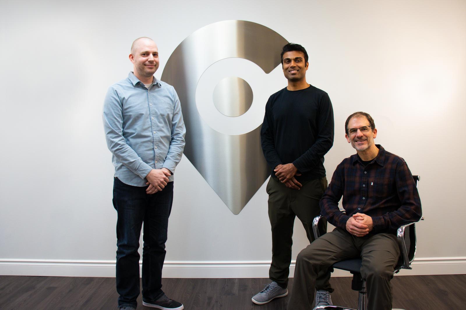 Prem Gururajan standing at the centre of a group of three RideCo colleagues in front of the company's location-pin emblem.