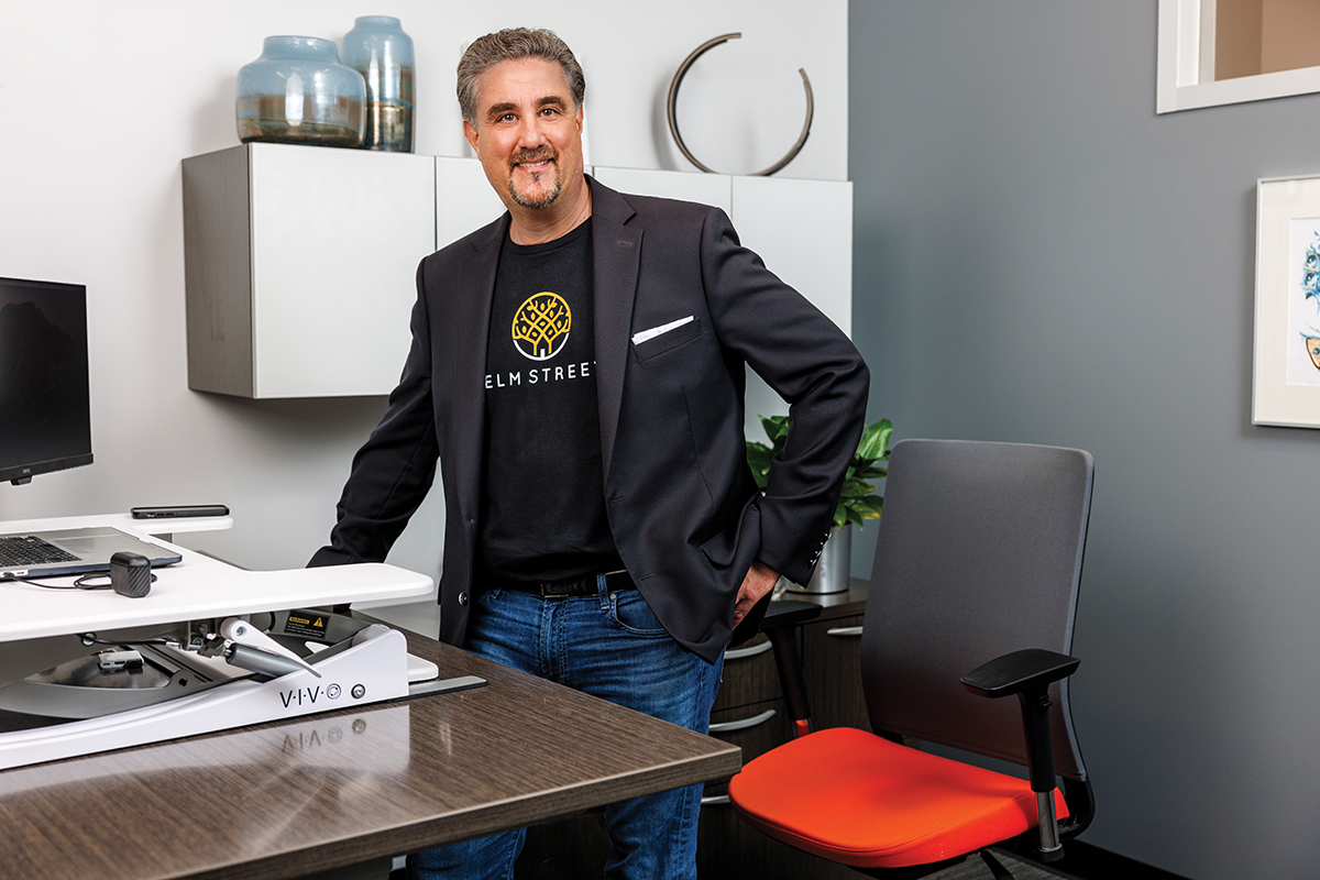 Prem Luthra standing beside a desk, wearing an Elm Street shirt and dark blazer.