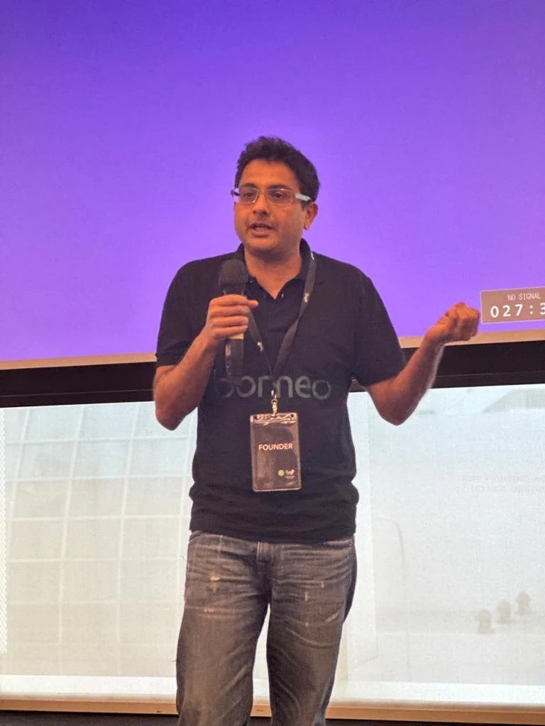 Prithvi Rai speaking with a handheld microphone, wearing a Borneo shirt and a founder badge.