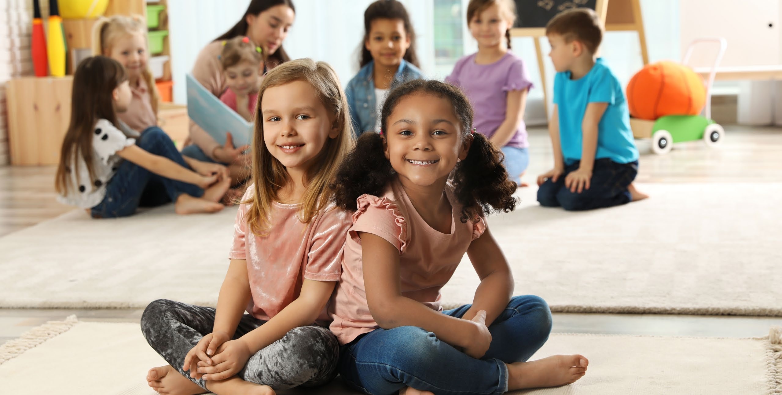 Children seated in a bright preschool classroom with a teacher reading behind them