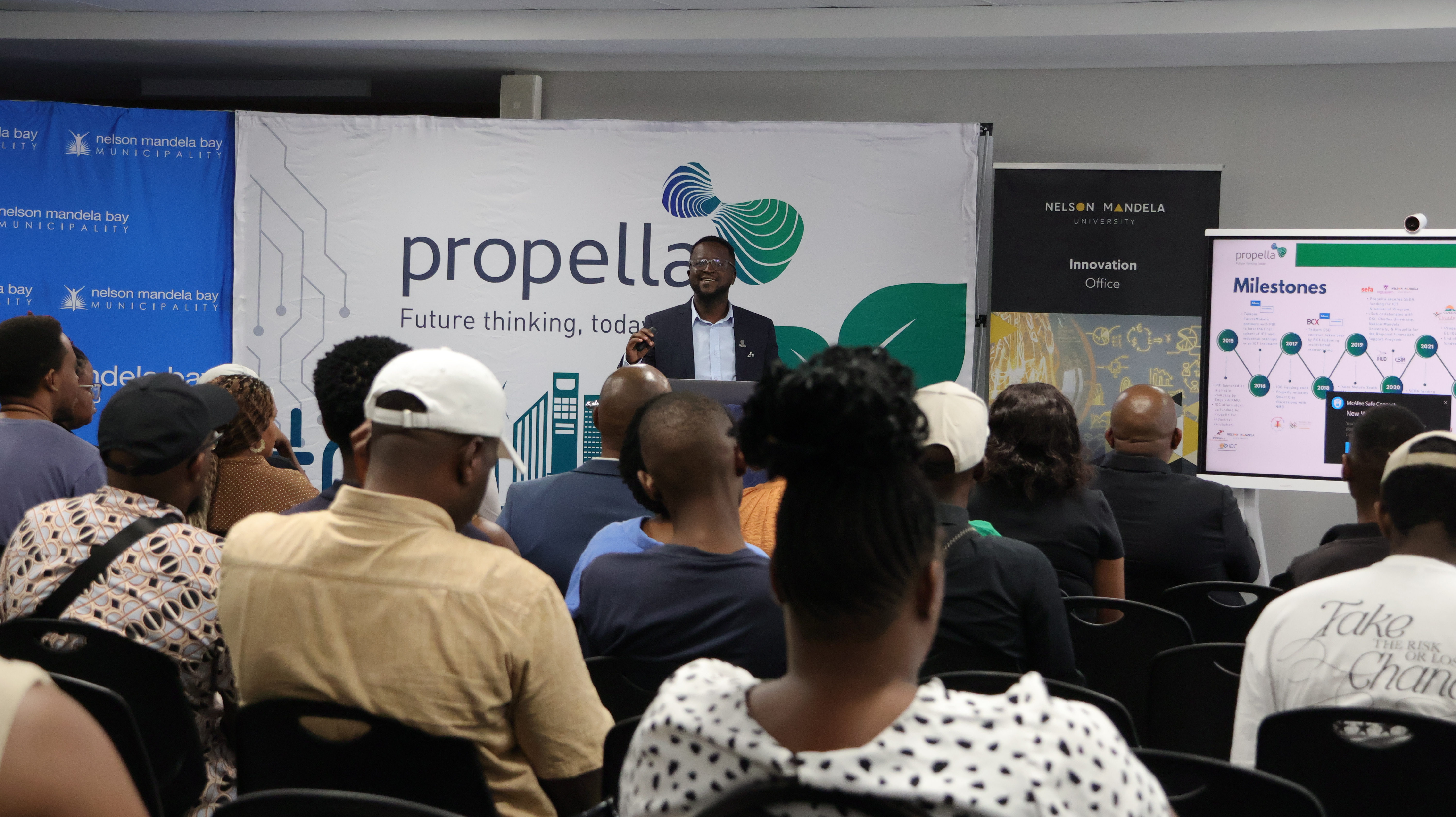 An audience listens to a speaker at a Propella gathering, with university and municipality banners behind him