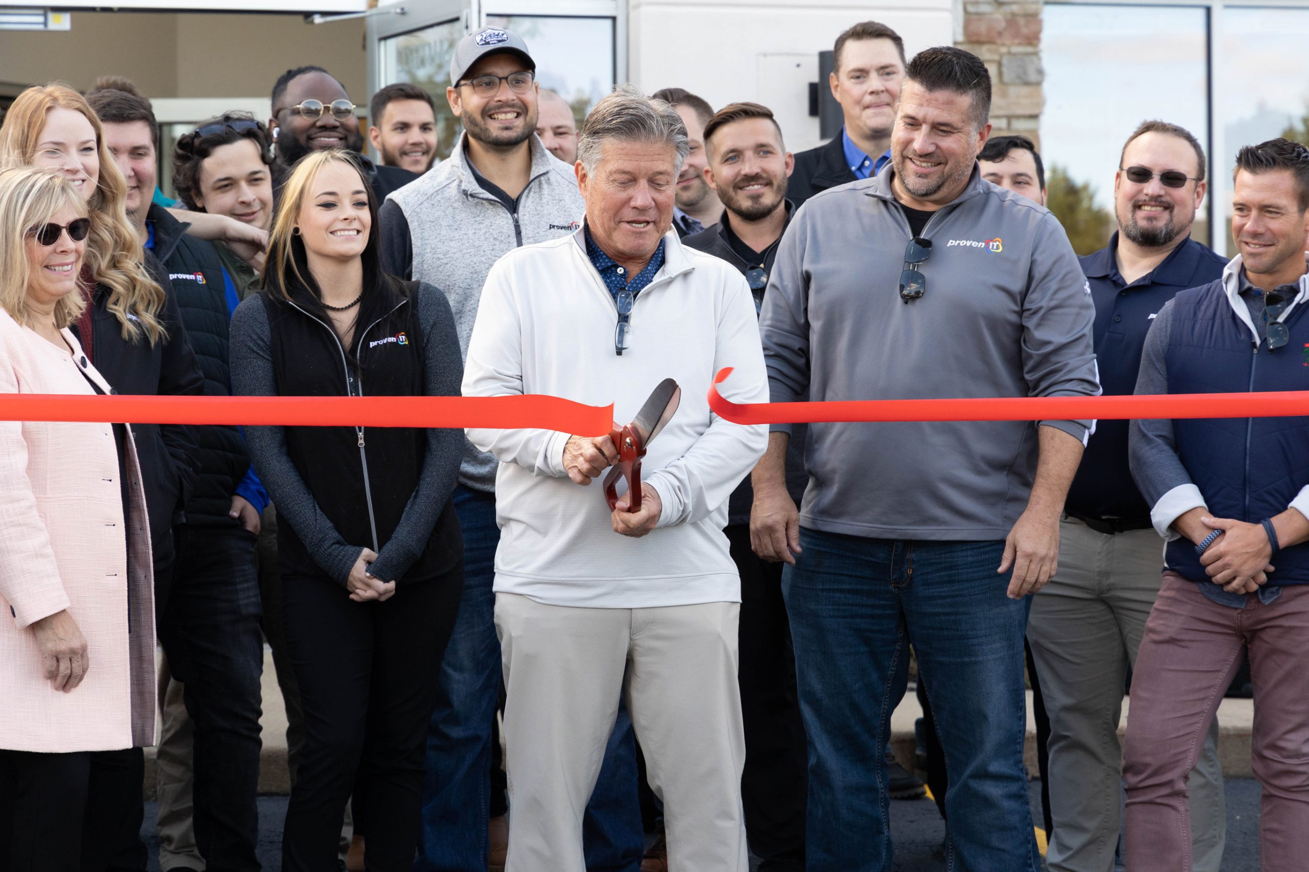 A group gathered around a red ribbon as a participant cuts it with large scissors