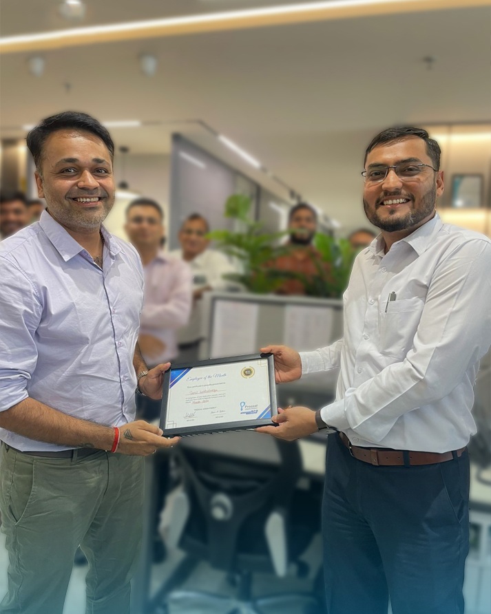 Two Prozeal colleagues holding a framed certificate in an office, with colleagues behind them