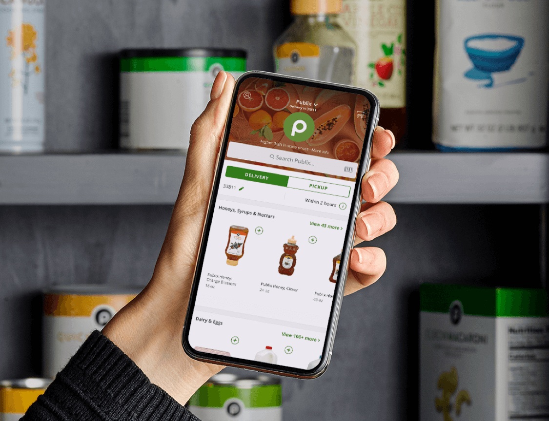 A shopper holds a phone showing Publix delivery and pickup ordering in front of pantry shelves