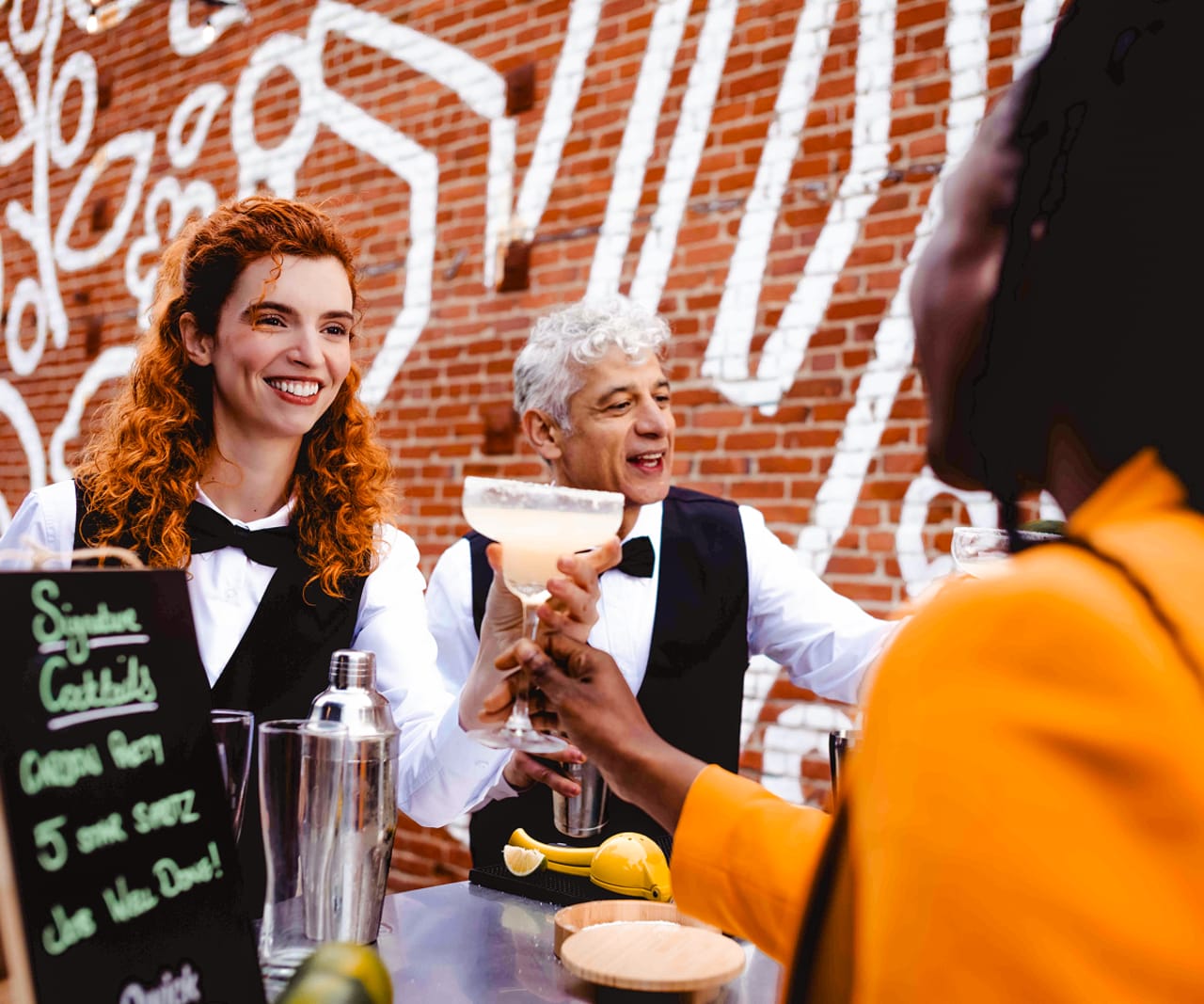 Two bartenders in vests and bow ties serving a coupe cocktail across a bar to a guest, with a Signature Cocktails chalkboard in the foreground