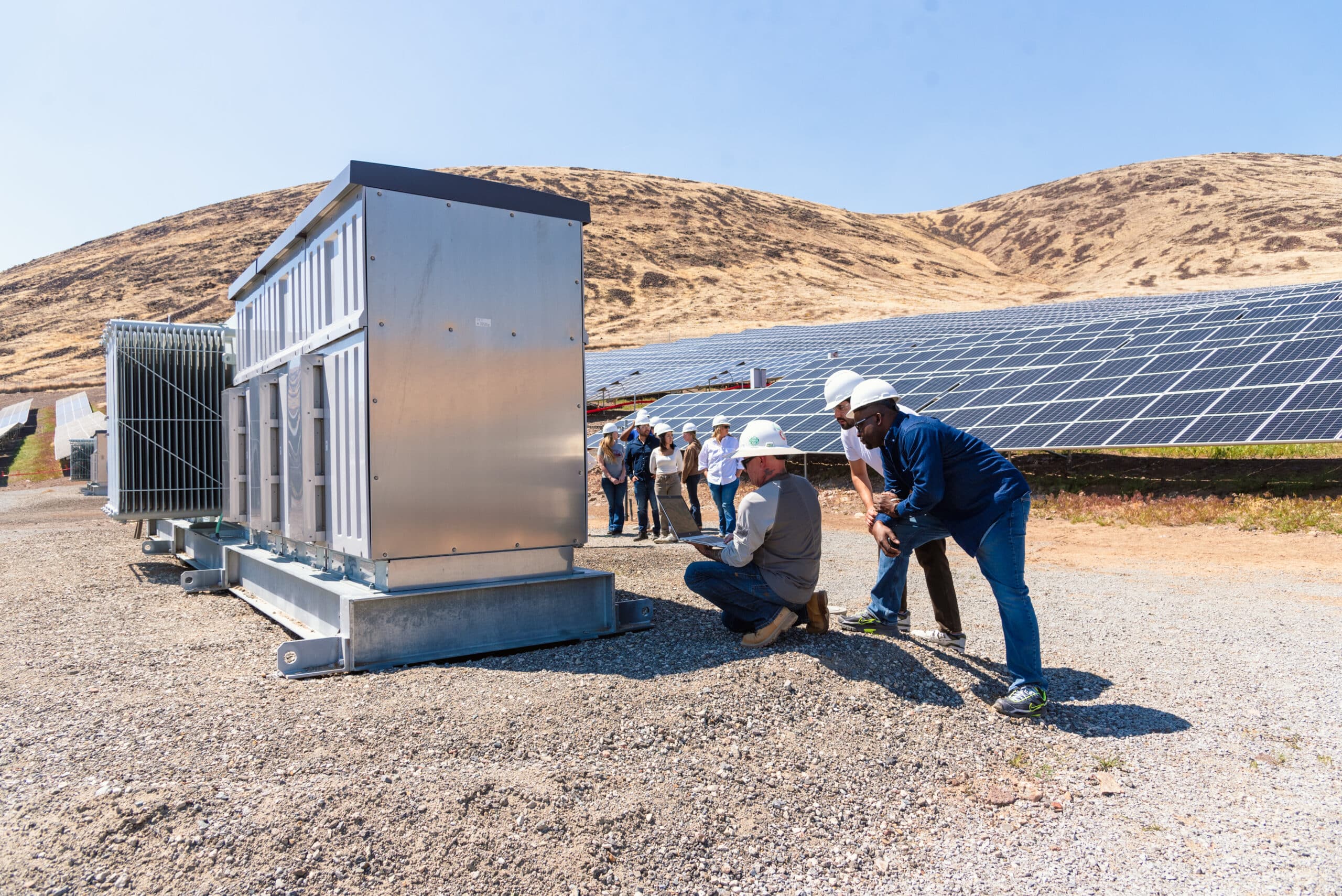 A group in hard hats examines electrical equipment beside rows of solar panels