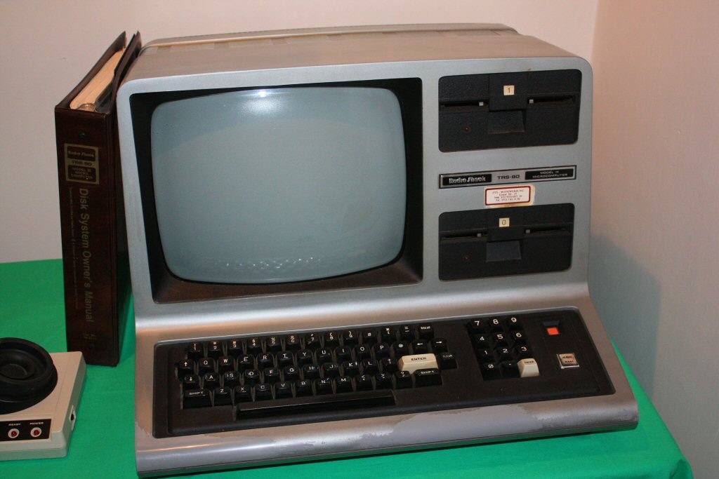 A silver RadioShack TRS-80 Model III computer with monitor, keyboard and twin disk drives
