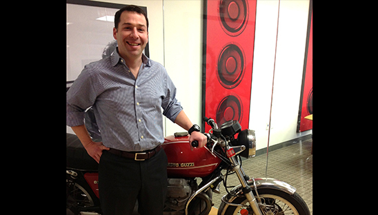 Rafi Musher standing beside his red Moto Guzzi motorcycle in an office