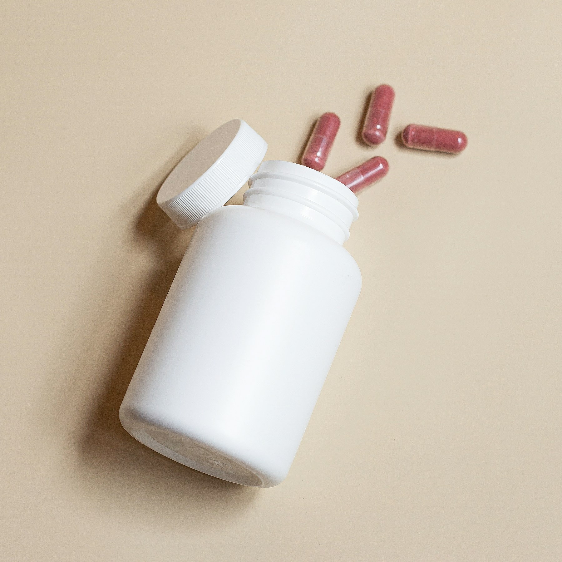 An open white supplement bottle and red capsules on a beige surface