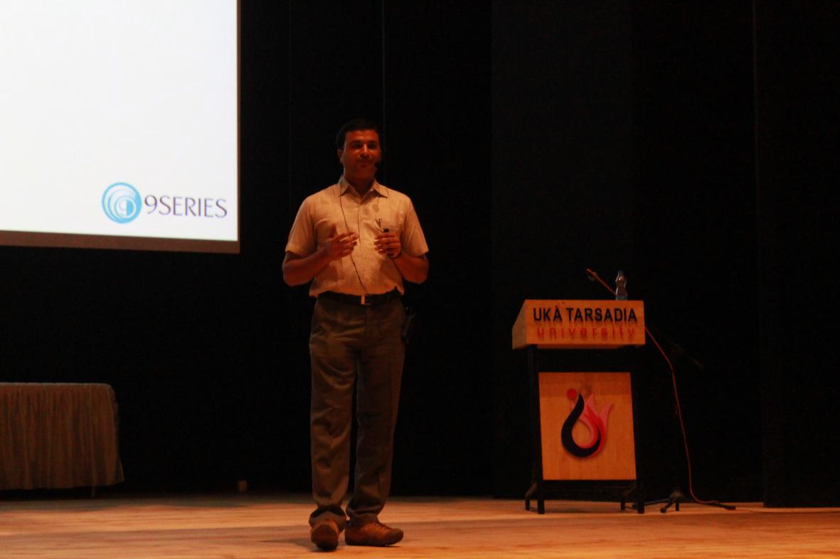 Rajnikant Joshi speaking beside a presentation screen at Uka Tarsadia University in 2015