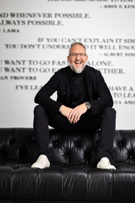 Randall Ward smiling while seated on a black leather couch, with quotations on the wall behind him