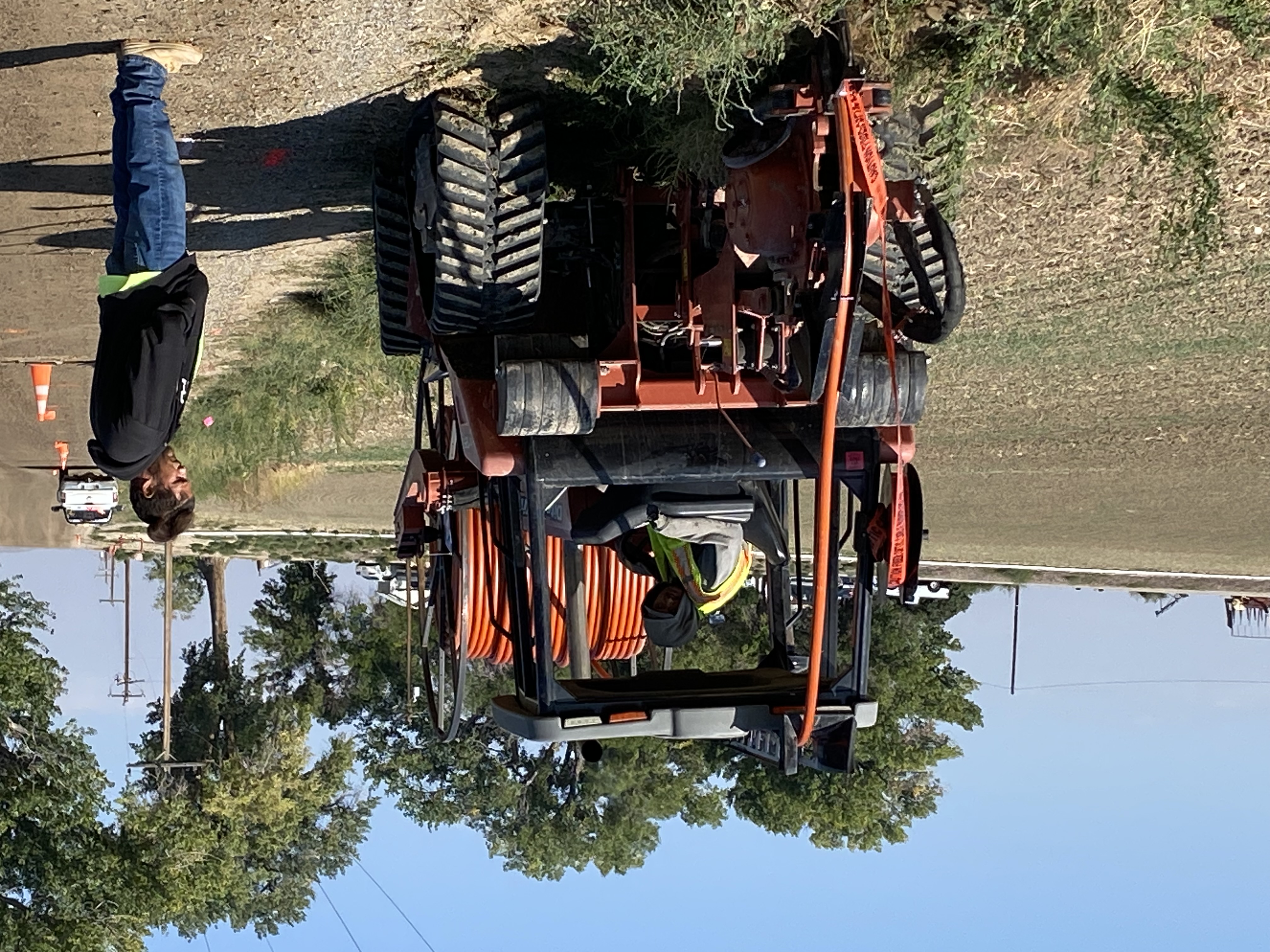 Workers beside a construction vehicle carrying orange conduit on a rural roadside