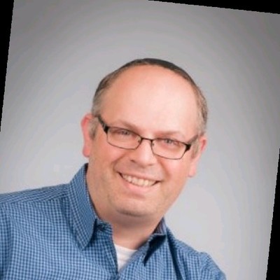 Raphael Nadel smiling, wearing glasses and a blue checked shirt