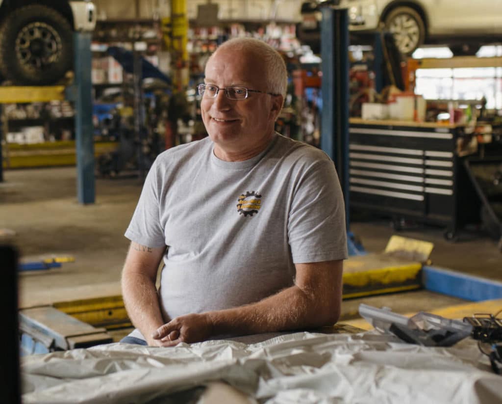 A Rapid Finance customer seated in an automotive workshop with vehicle lifts and tools behind him