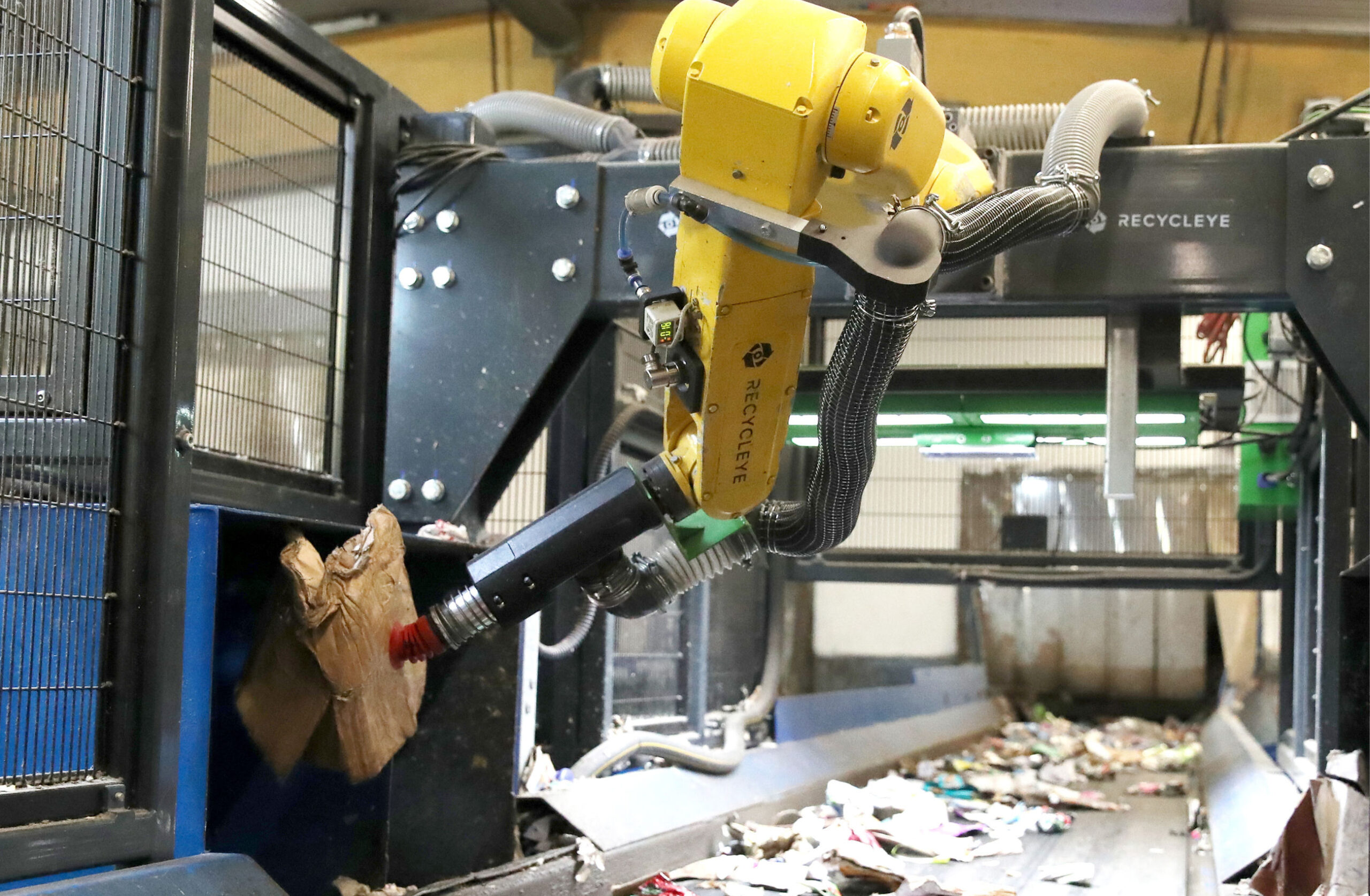 Yellow Recycleye robotic arm lifting a piece of cardboard beside a recycling conveyor