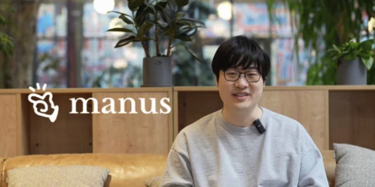 Red Xiao wearing glasses and a grey sweatshirt, seated during an interview in front of books and plants.