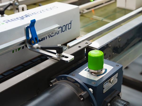 A green condition-monitoring sensor mounted on a conveyor gearbox beside an electric motor