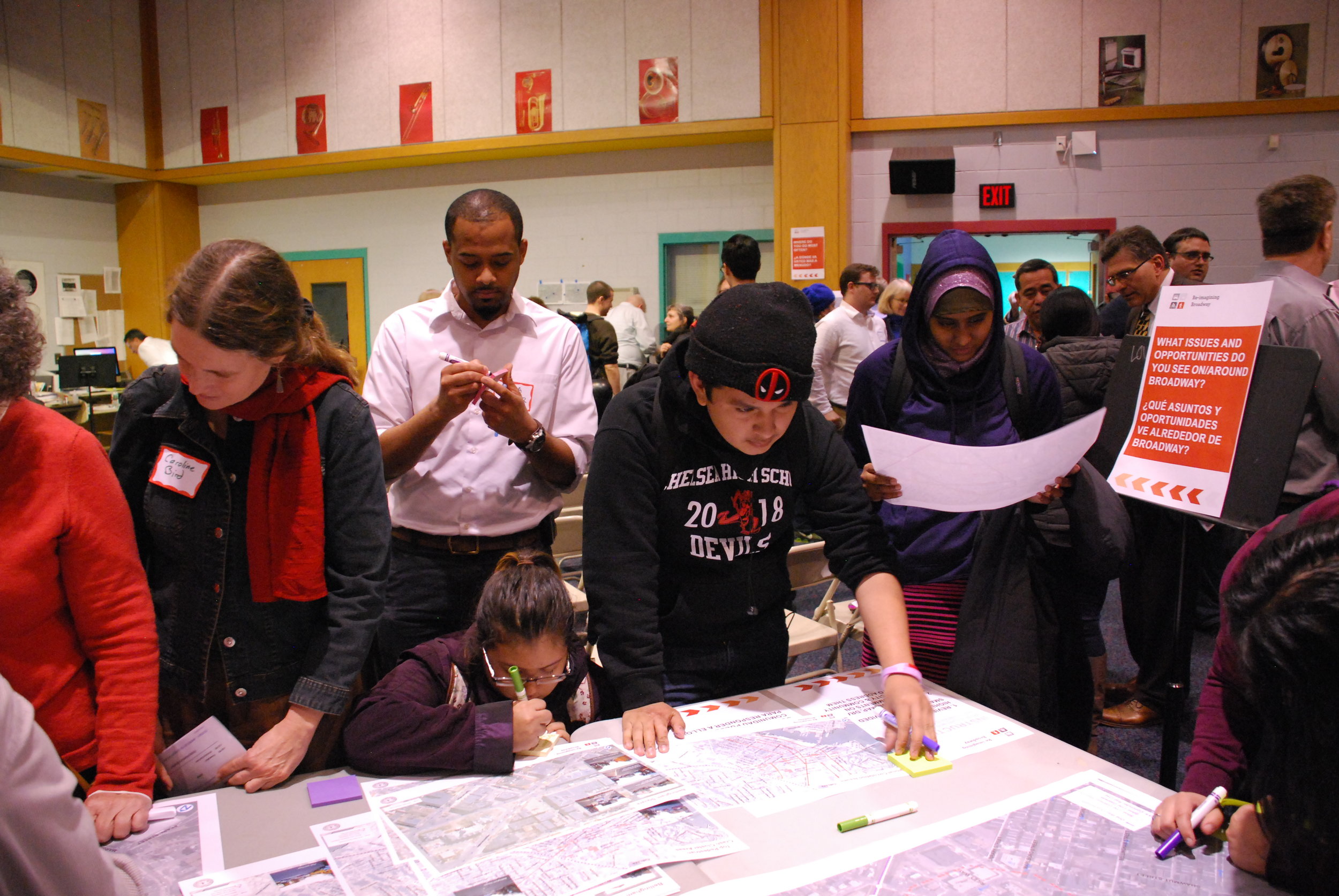 Residents at a crowded public meeting write comments on maps while a bilingual sign asks about Broadway