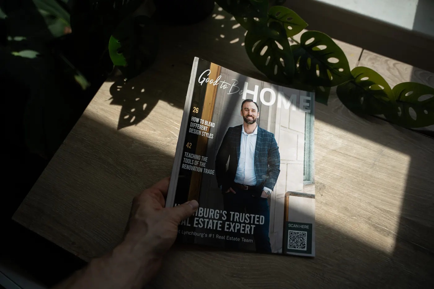 A hand holding a personally branded Good to Be Home magazine on a sunlit wooden table