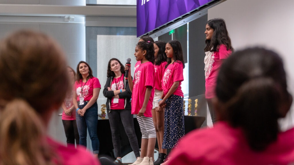 Girls in pink GLAM shirts presenting together on a stage while an audience watches