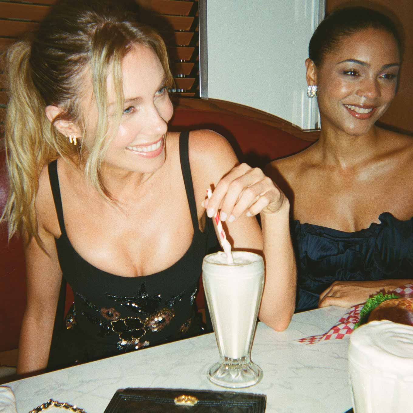 Two women wearing evening outfits at a diner table with a milkshake