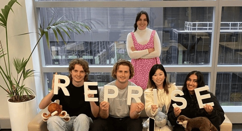 Five people in a Reprise office, four seated holding letters spelling Reprise and one standing behind the sofa.