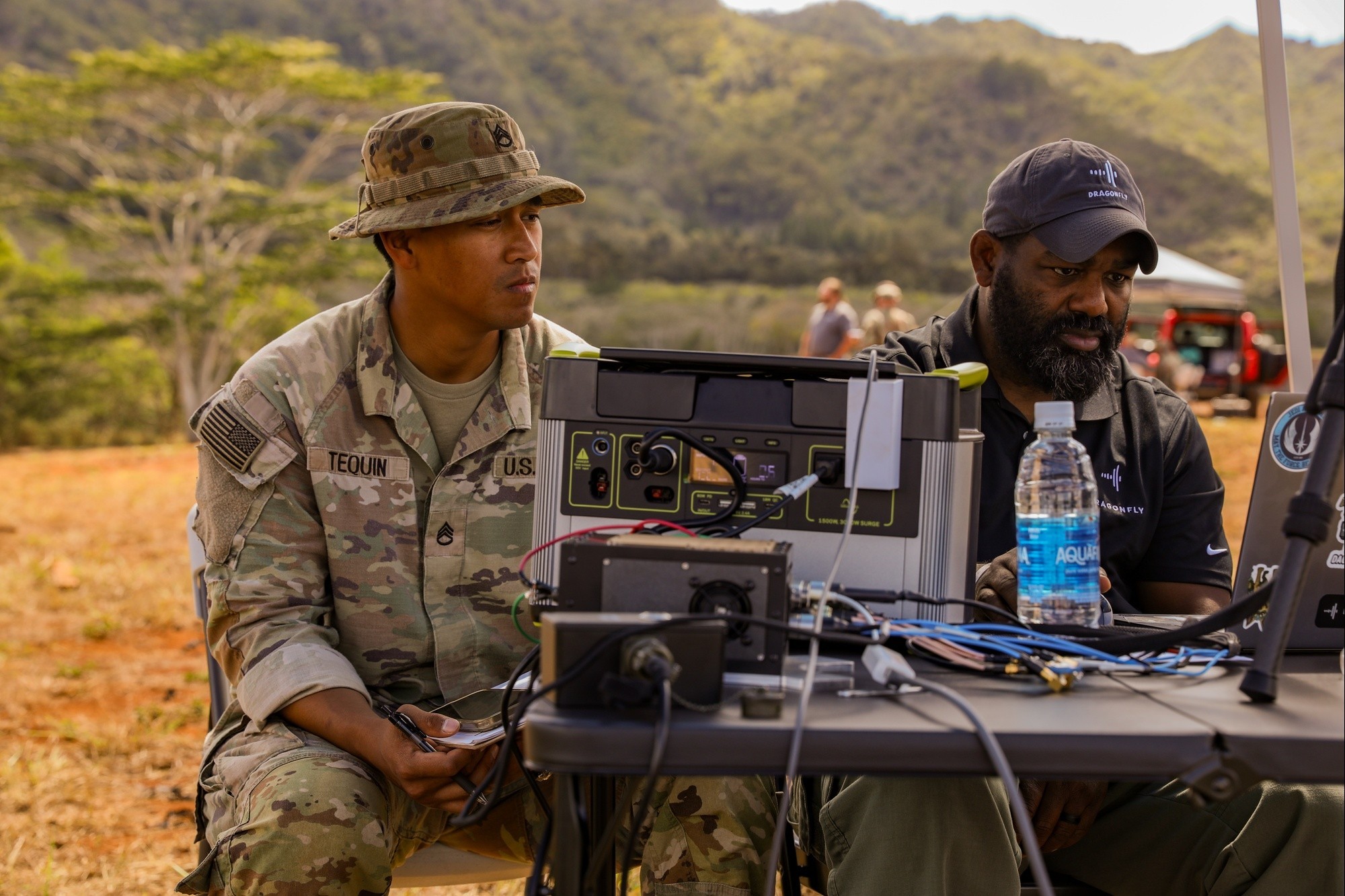 A U.S. soldier and a Dragonfly representative study a laptop beside signal-analysis equipment outdoors