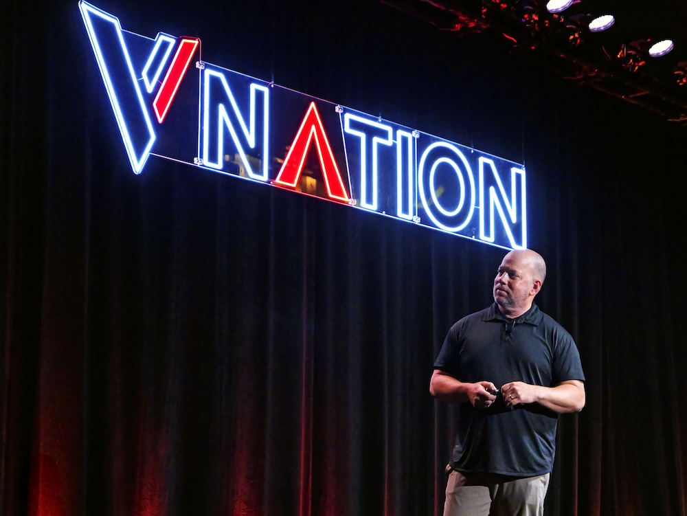 Rich Balot speaking on stage underneath a lit VNATION sign