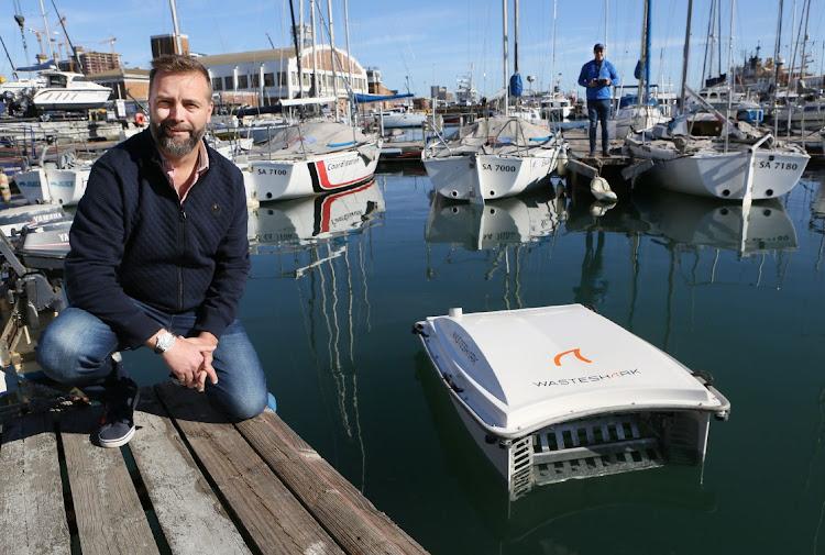 Richard Hardiman crouching on a Cape Town dock beside a WasteShark, with moored sailing boats behind him