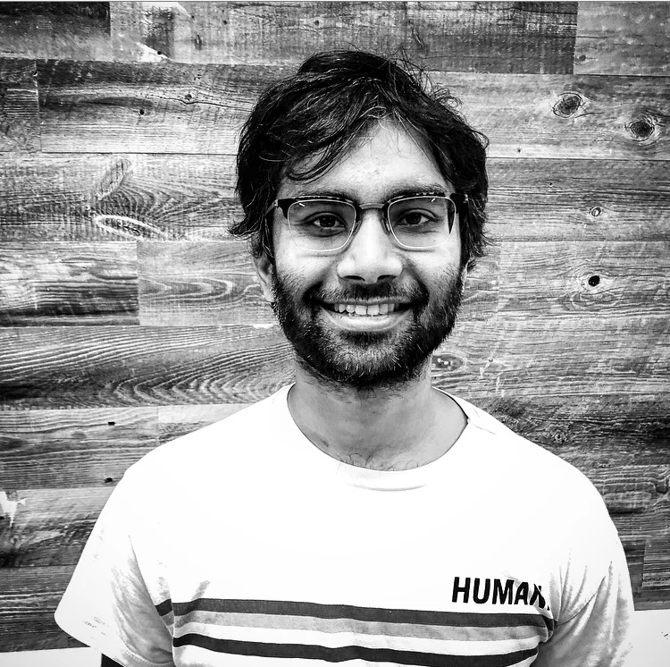 Ricky Sahu smiling in a black-and-white photograph by Mike Jabbawy, wearing glasses and a shirt marked HUMAN.