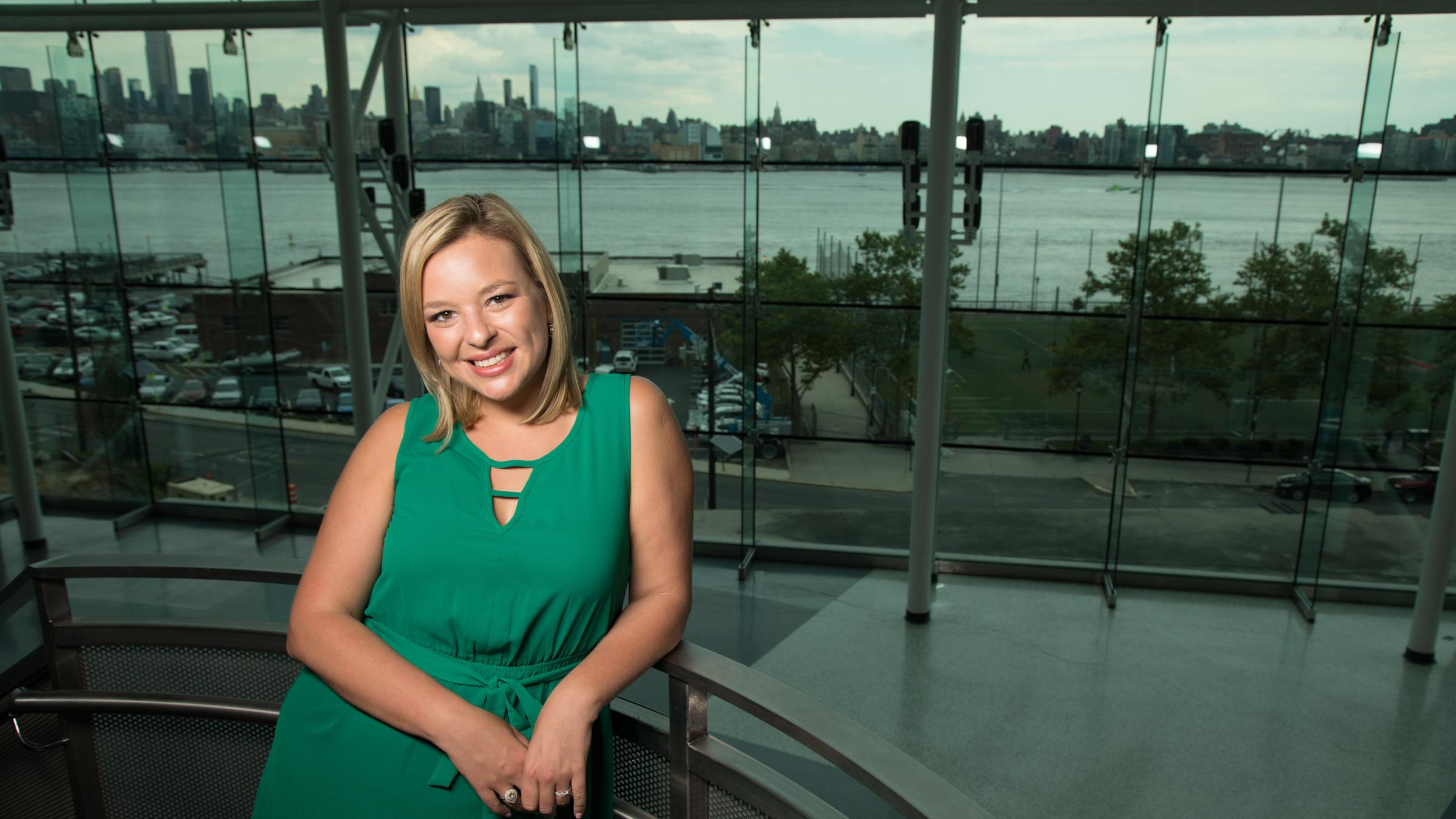Rita Gurevich seated inside a glass-walled building with the Manhattan skyline across the river