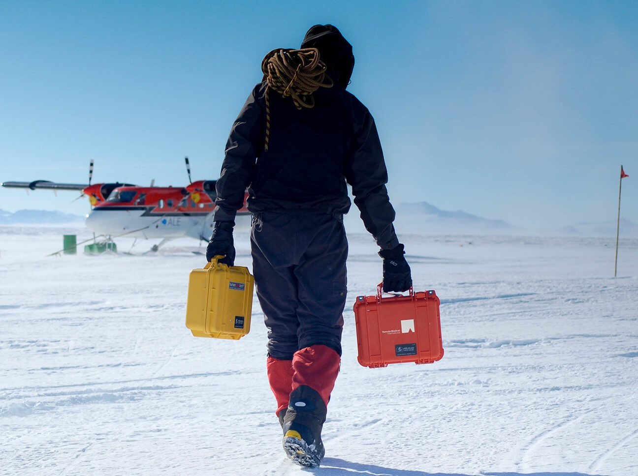 A person carries yellow and red equipment cases across snow toward a waiting aircraft