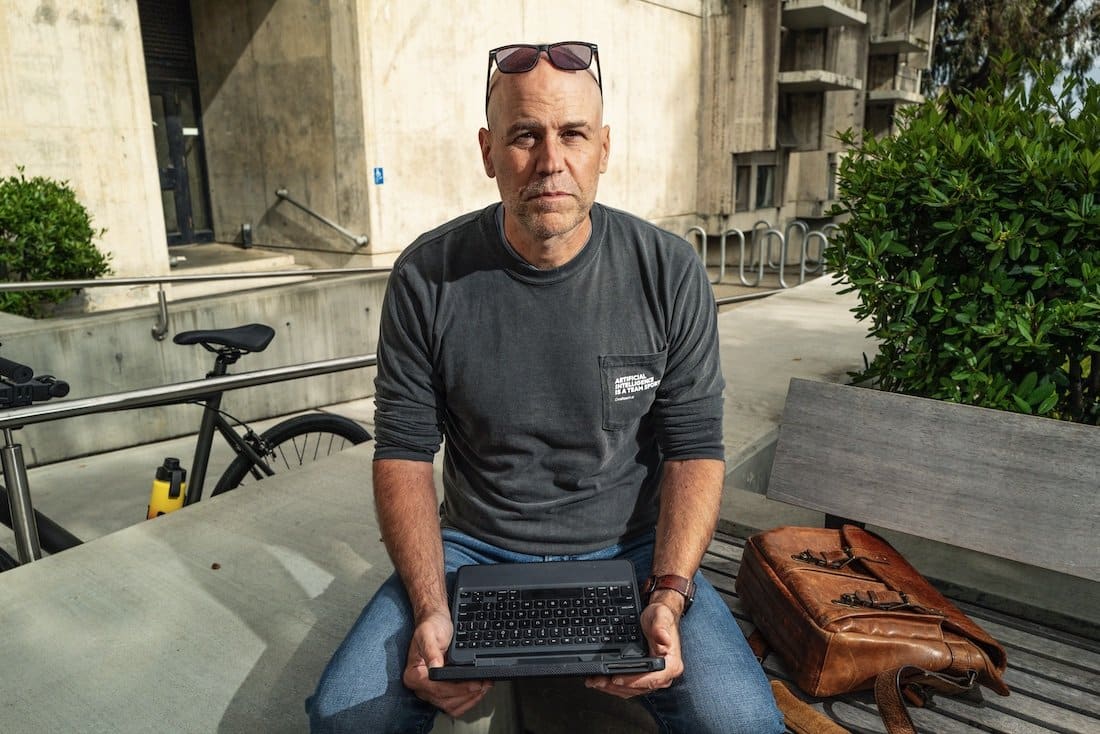 Robb Wilson seated outdoors with a small computer on his lap and a bicycle nearby