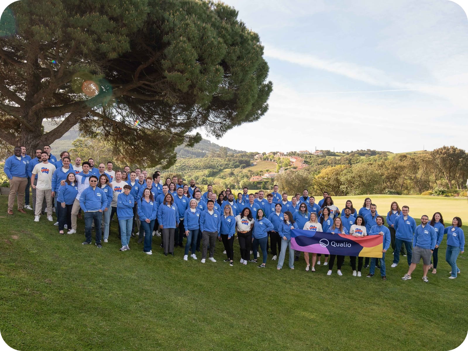 Qualio colleagues gathered outdoors beneath a large tree, many wearing blue company sweatshirts
