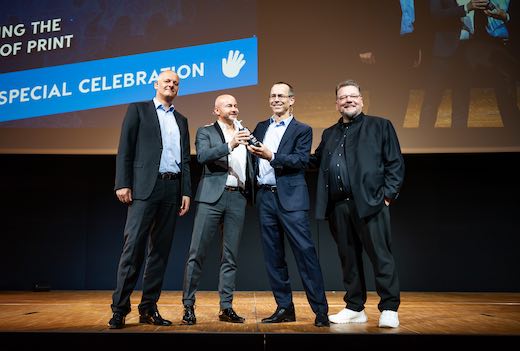 Four men on stage at the 2023 Online Print Symposium; Robert Keane, second from right, receives an award from Thorsten Fischer.