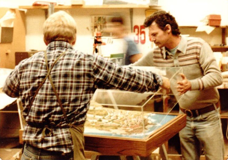 Archival MSI workshop photograph showing employees handling a clear cover above an architectural scale model