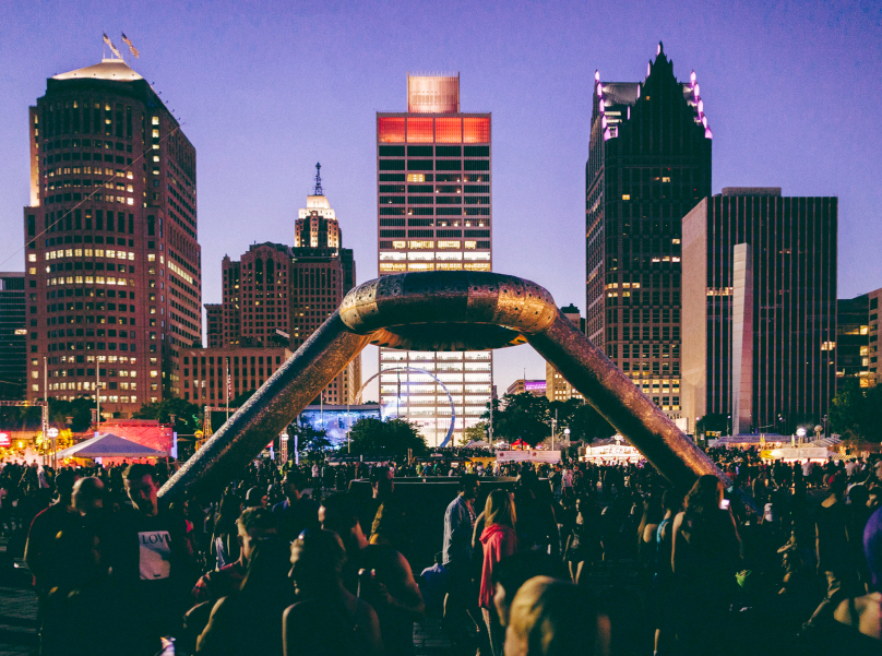 A crowd gathers in downtown Detroit beneath a large illuminated arch, with office towers behind.