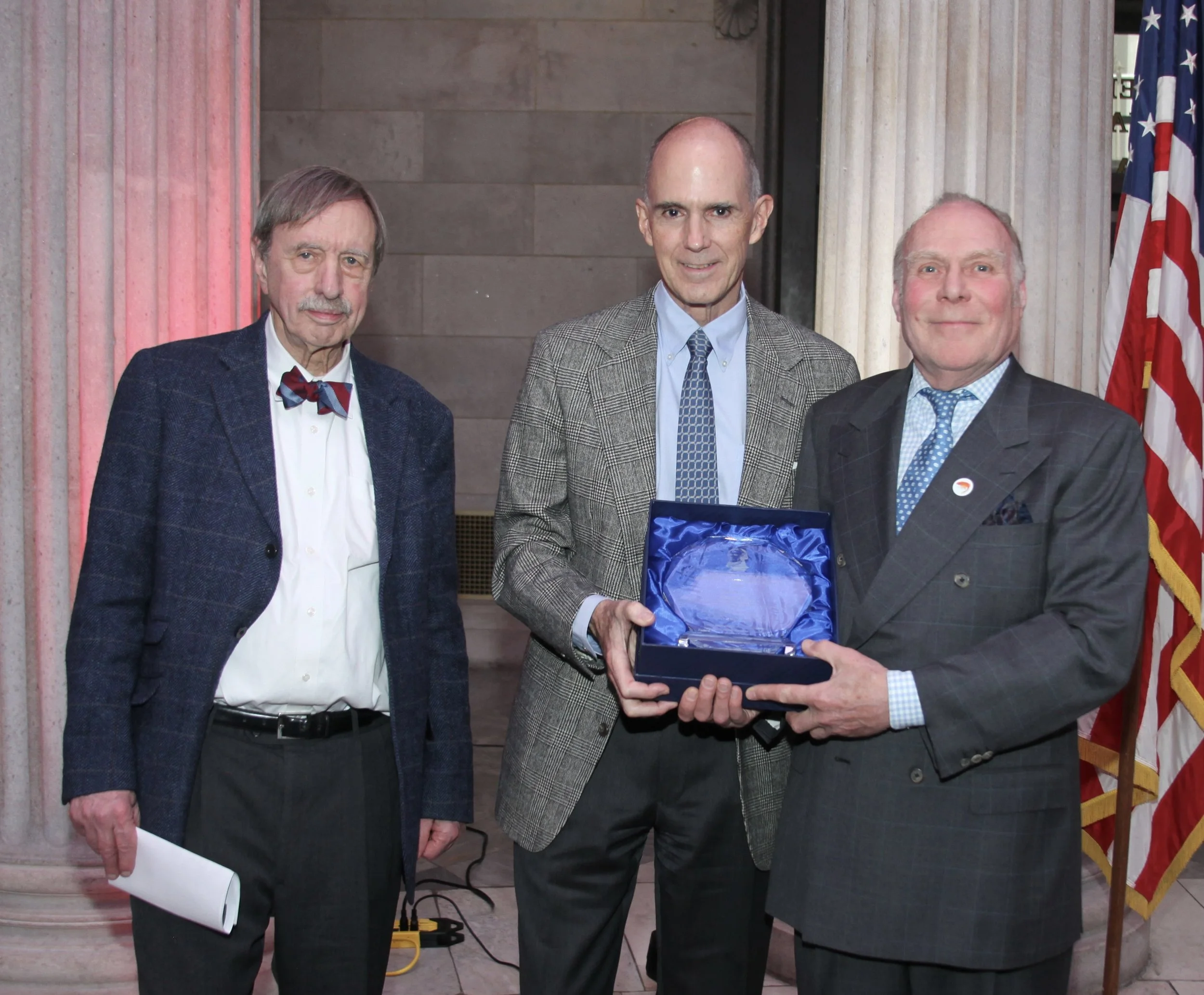 Roger Byrom holding his award between two presenters at the 2026 immigrant achievement ceremony