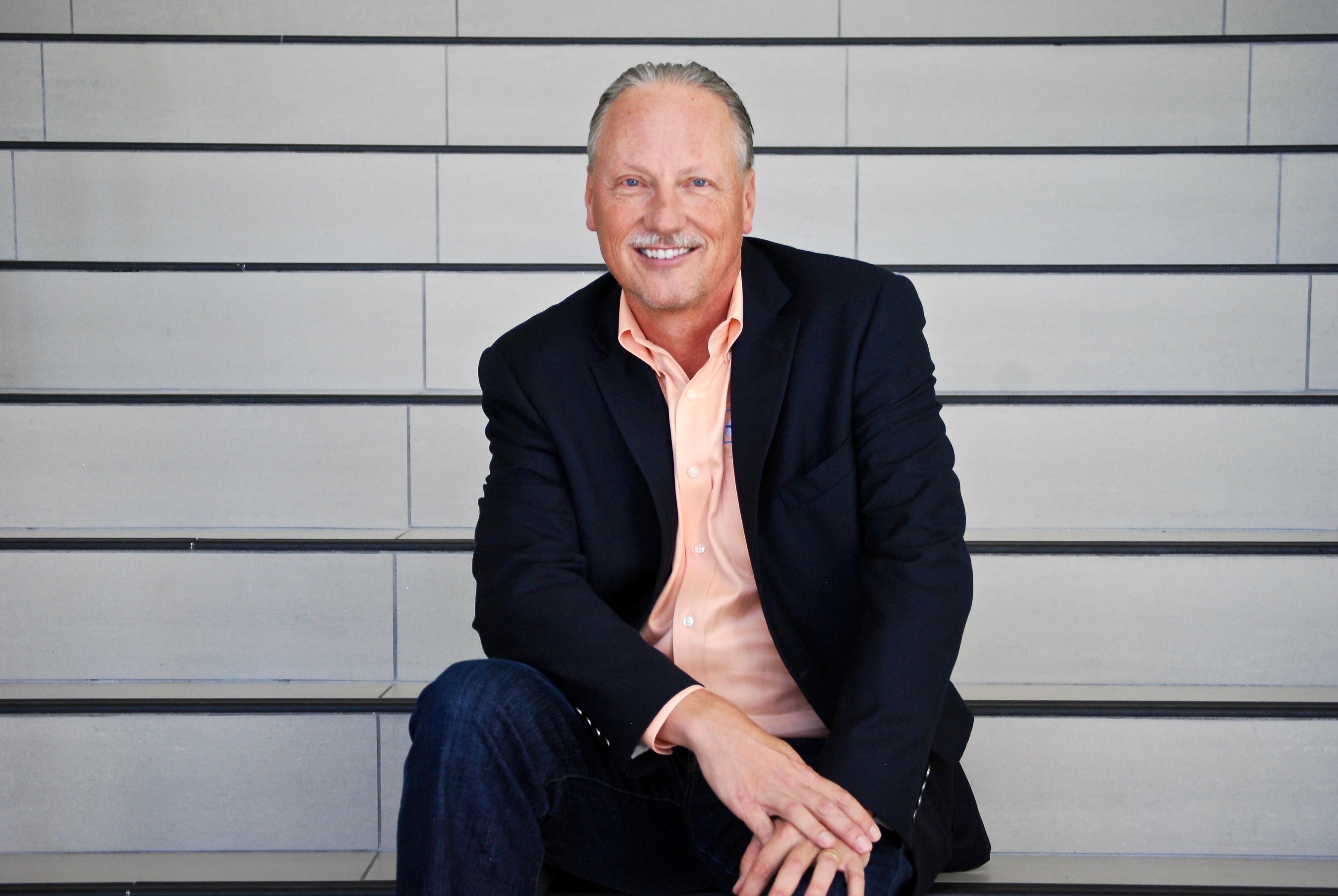 Ron Daly seated on wide concrete steps, wearing a dark blazer and coral shirt