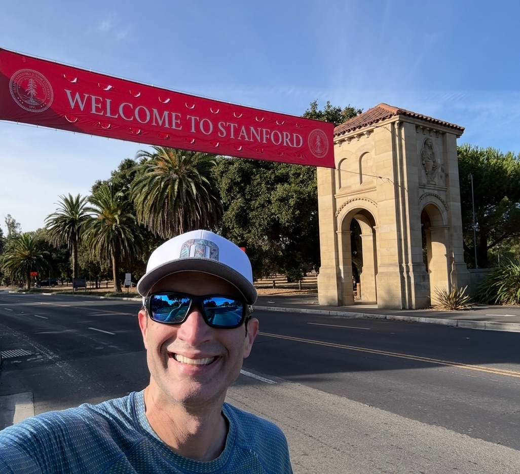 Ron Gutman taking a selfie beside Stanford University's entrance and welcome banner