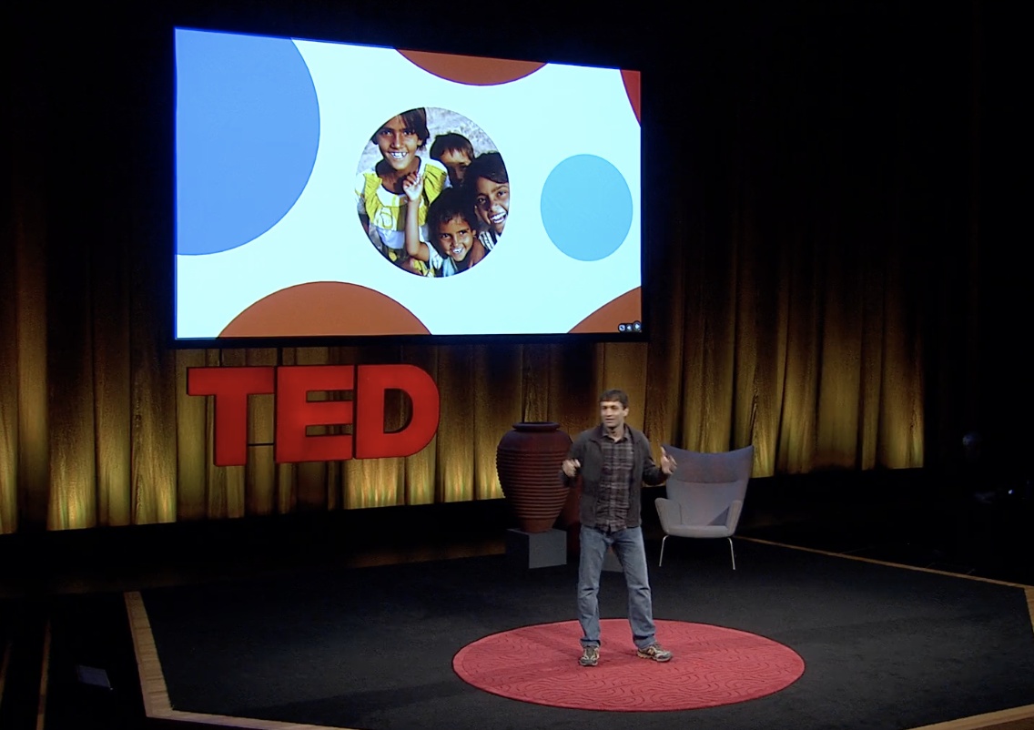 Ron Gutman presenting on the TED stage, with a photograph of smiling children on the screen