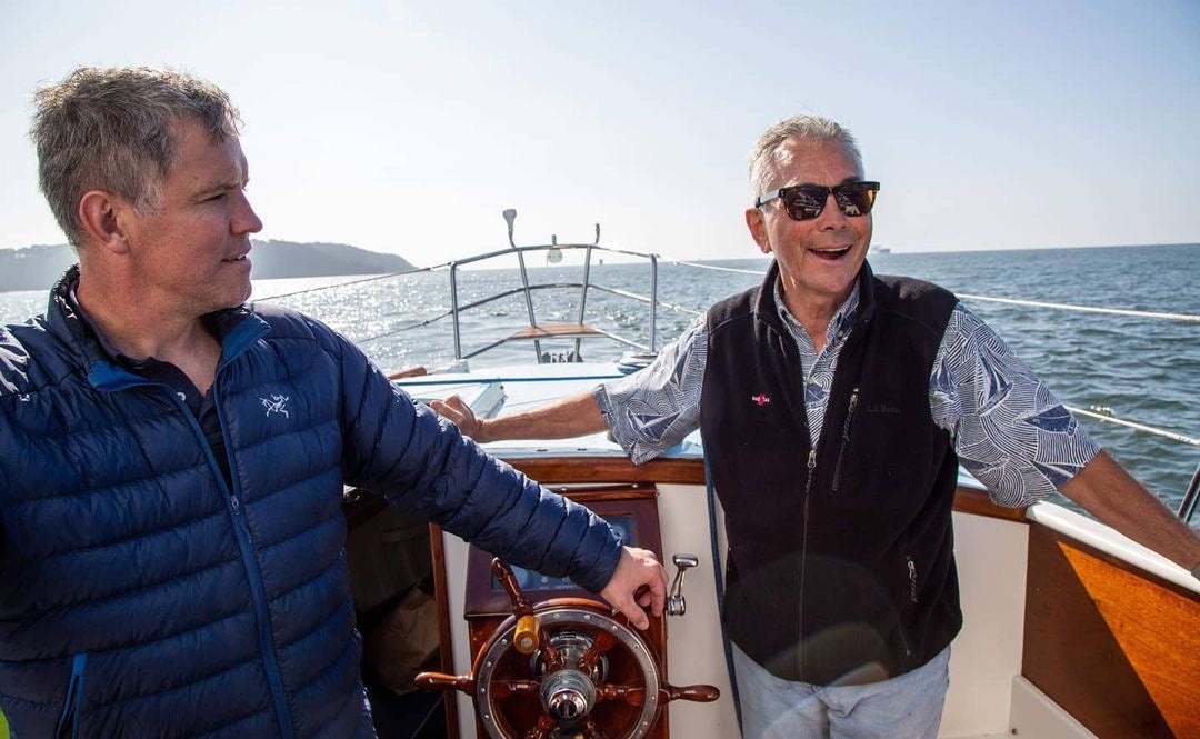 Ron Young, at right in sunglasses, smiles aboard a sailboat beside another sailor.