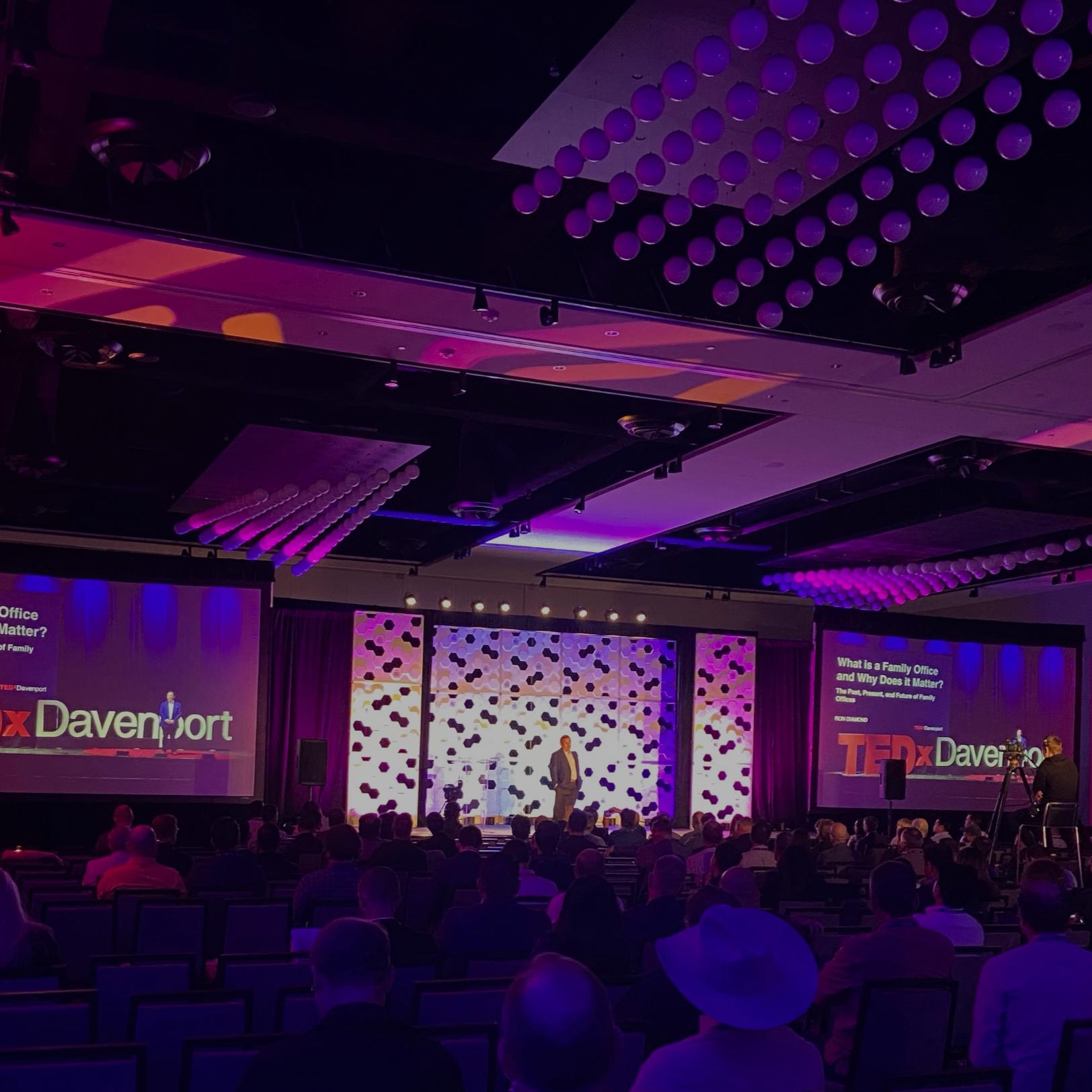 Ronald Diamond on the TEDxDavenport stage, seen from the seated audience beneath purple auditorium lighting