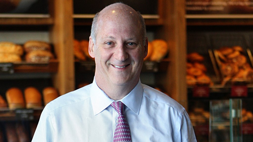 Ron Shaich standing in a bakery café, with shelves of bread behind him