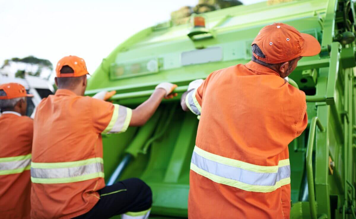 Waste collection workers in orange uniforms riding on a green refuse truck